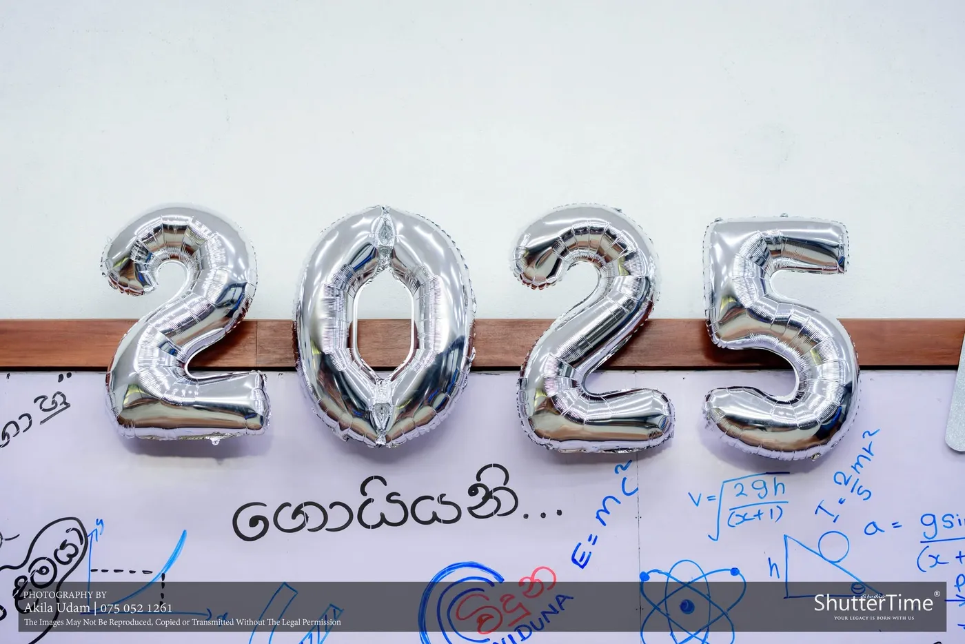 රිදී පැහැති 2025 බැලූන් අකුරු, පසුබිමේ ලියූ වයිට් බෝඩ් එකක්