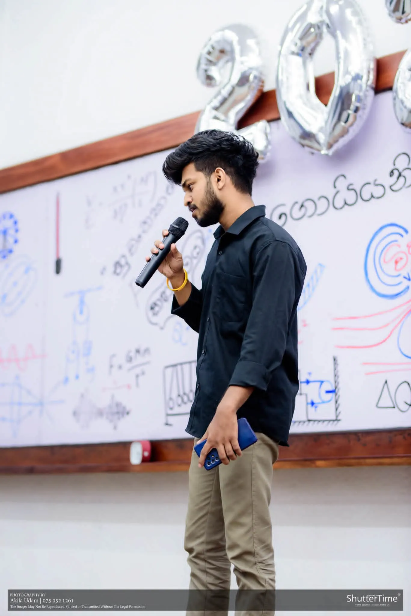 සිසුවෙක් වයිට් බෝඩ් එකක් ඉදිරිපිට මයික්‍රොෆෝනයක් අතැතිව