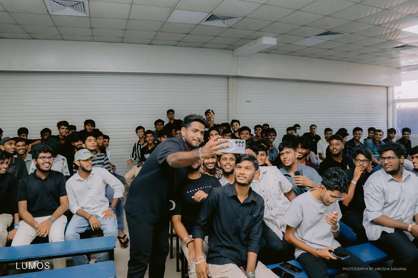 ශාලාවේ සිසුන් සමූහයක්, ඉදිරියෙන් අයෙක් අත දිගු කර